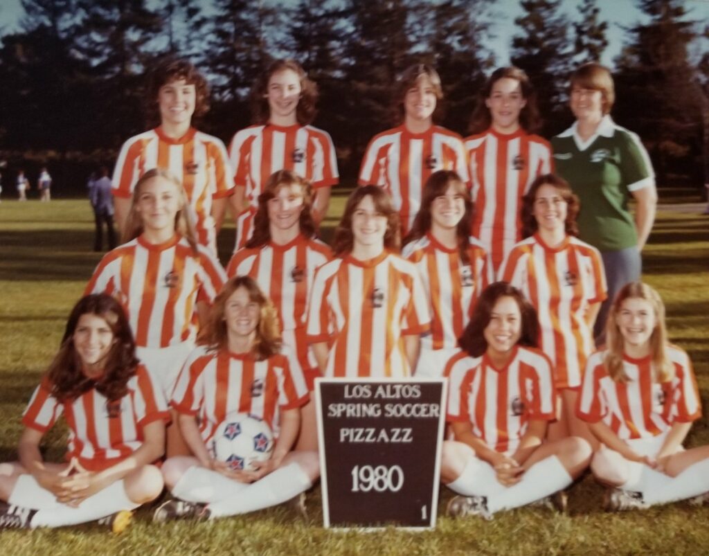 A girls’ soccer team poses outdoors in orange and white striped uniforms, smiling with their coach. A sign in front reads: Los Altos Spring Soccer Pizza ZZ 1980. Trees and other players are visible in the background.