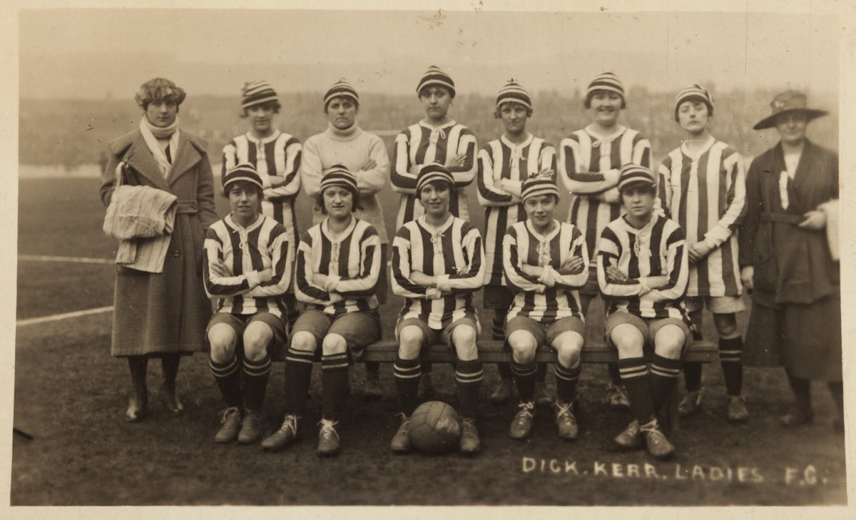 A vintage black-and-white photo of the Dick, Kerr Ladies F.C. womens football team, wearing striped jerseys and hats, posing on a football field with two women in coats standing at each end.
