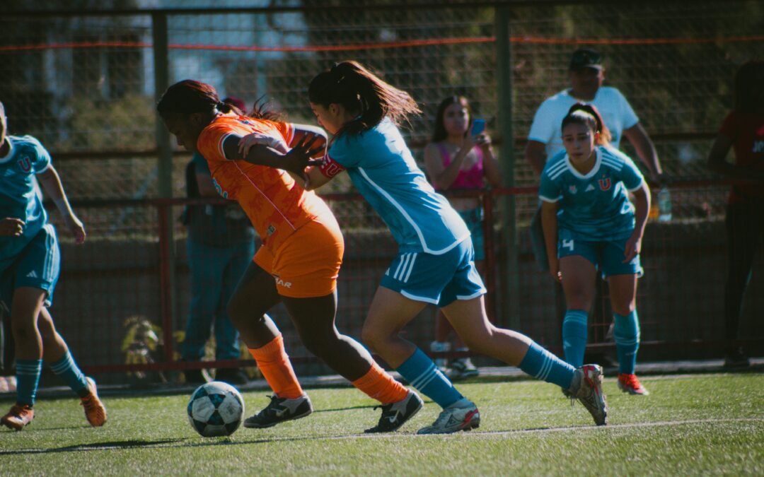 Two female soccer players compete for the ball on a field, one in an orange uniform and the other in blue. Other players and spectators are visible in the background behind a fence.